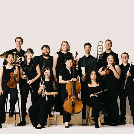 Twelve+musicians+in+black+pose+with+their+instruments+against+a+light+background%2C+smiling+at+the+camera.
