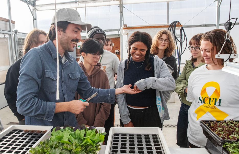 A farmer hands a student a cutting of a vegetable inside the greenhouse at Foothills Farm