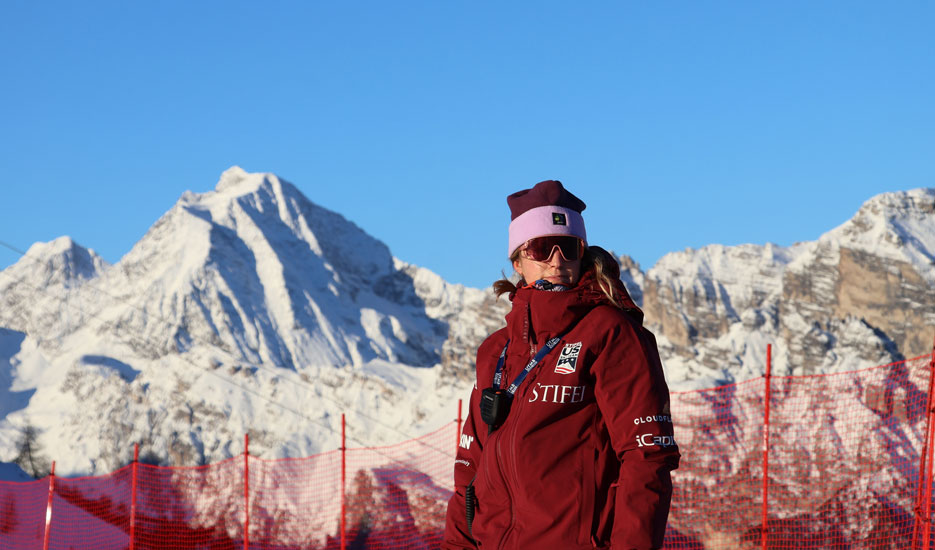 Lyndsay Strange '09 in a maroon ski jacket and pink hat standing in front of snow-covered mountain peaks and red safety fencing under a clear blue sky.