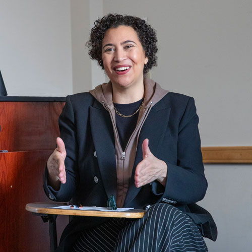 Meredith+Broussard+sits+at+a+classroom+desk%2C+smiling+and+gesturing+with+her+hands+as+she+speaks+to+students.