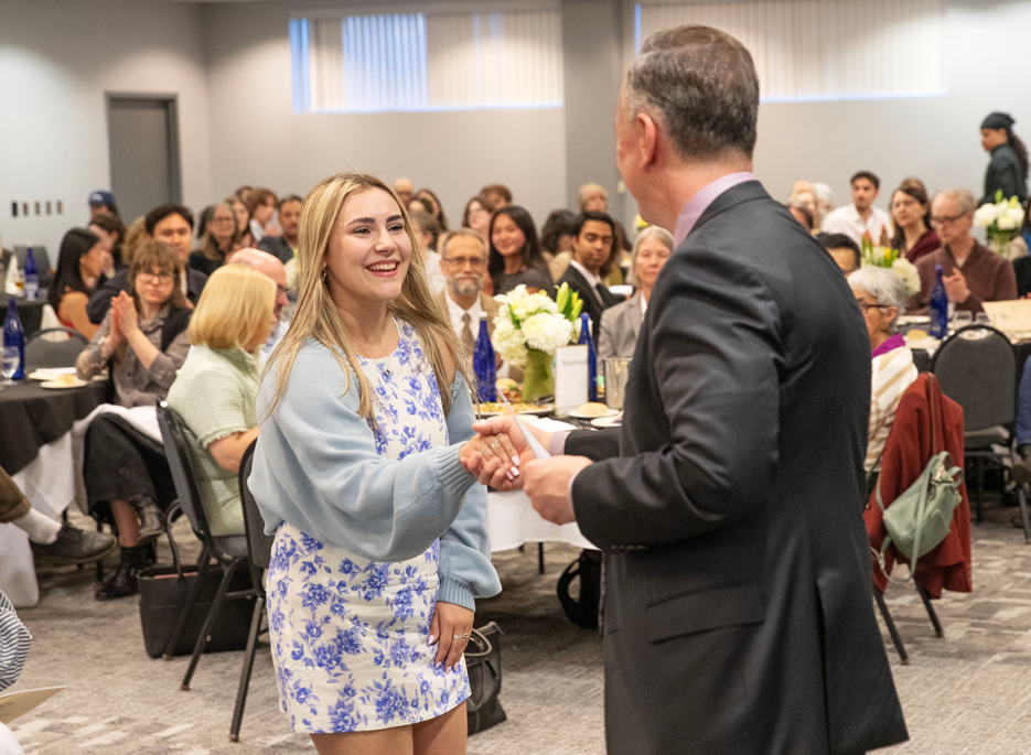 A student smiles and shakes the hand of Skidmore President Marc Conner as a room full of people looks on