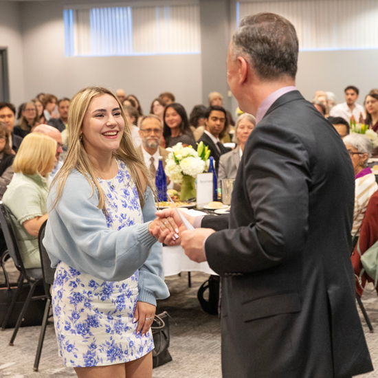 A+student+smiles+and+shakes+the+hand+of+Skidmore+President+Marc+Conner+as+a+room+full+of+people+looks+on