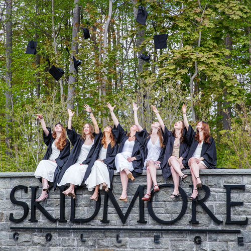 A+group+of+graduates+in+caps+and+gowns+celebrate+by+tossing+their+graduation+caps+into+the+air+while+sitting+on+a+stone+sign+in+front+of+a+wooded+backdrop+at+Skidmore+College.