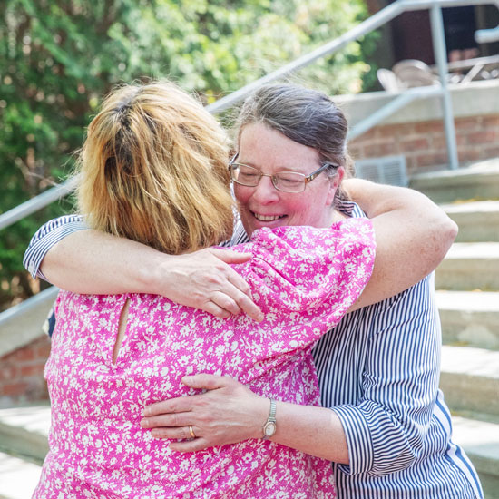 Two+colleagues+embrace+warmly+outside+near+a+campus+stairway%2C+smiling+as+they+hug+during+a+celebration.