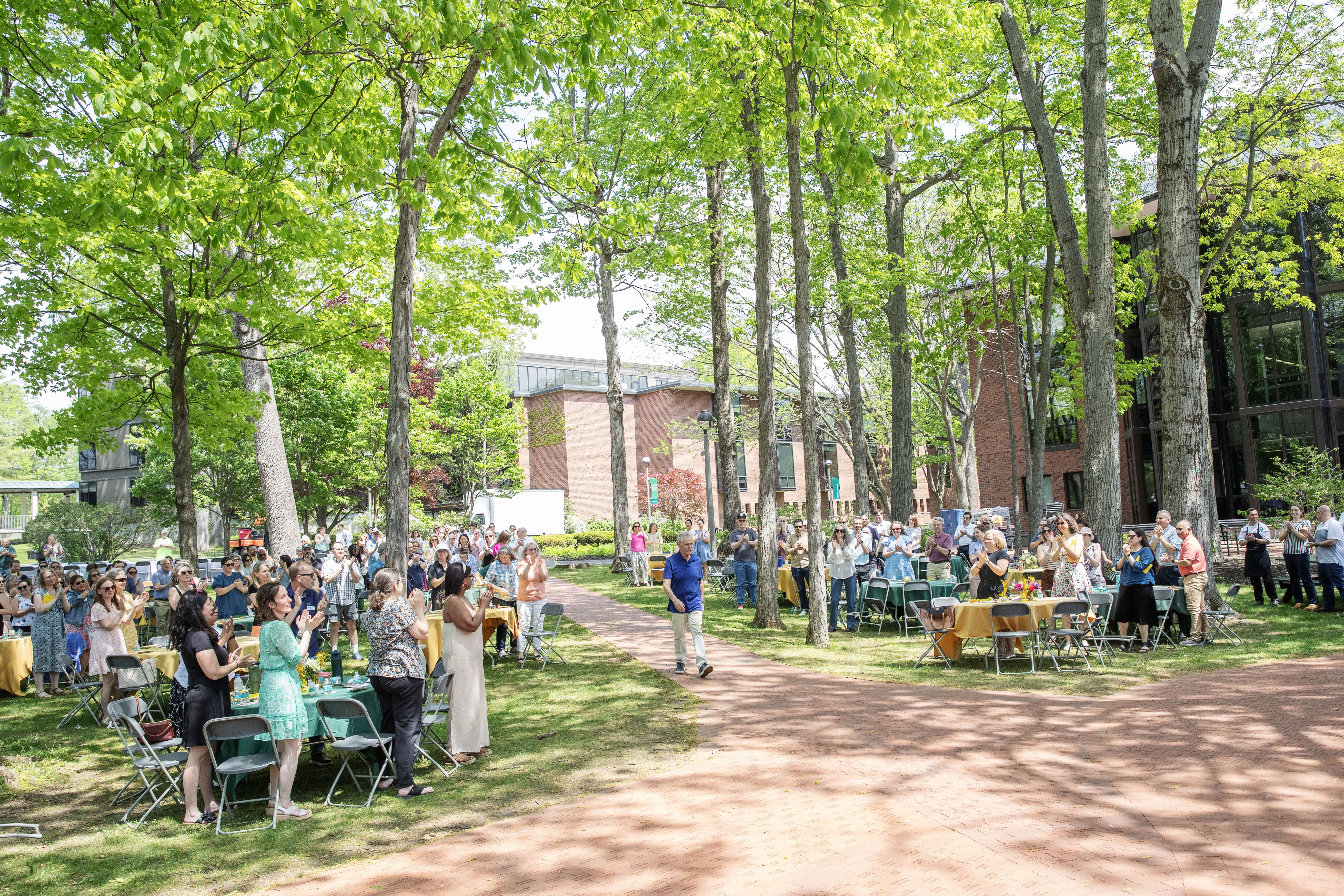 A large crowd of colleagues stands and applauds as Daniel Hurwitz walks along a brick pathway through a campus gathering beneath tall green trees.