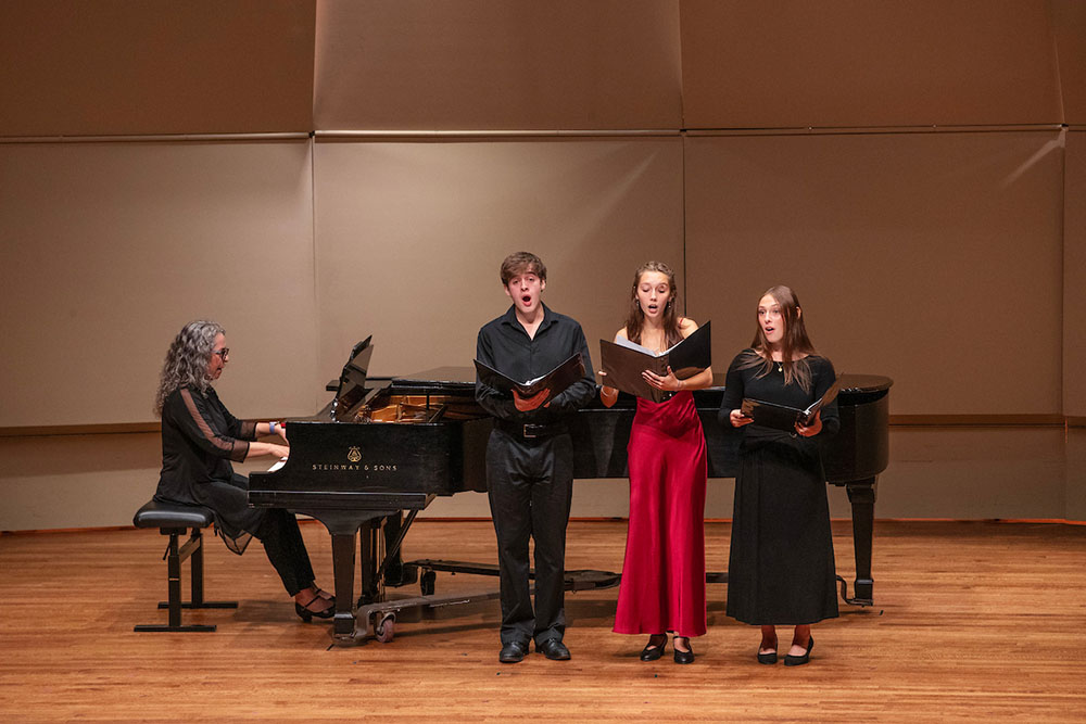Three students stand in front of grand piano singing during a performance on stage, one person sits at piano bench playing.