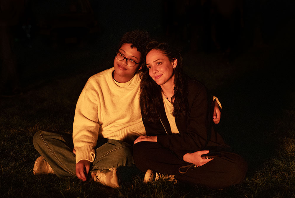 Two people sit on the ground next to each other in front of a bonfire. 
