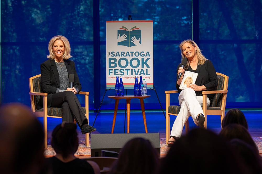 Two people sit in chairs on a stage for a talk with a poster on an easel in between them. One is holding a microphone. 