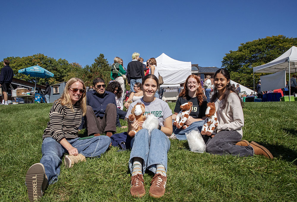 Five people sit on the grass, smiling, at a artisan craft festival. 