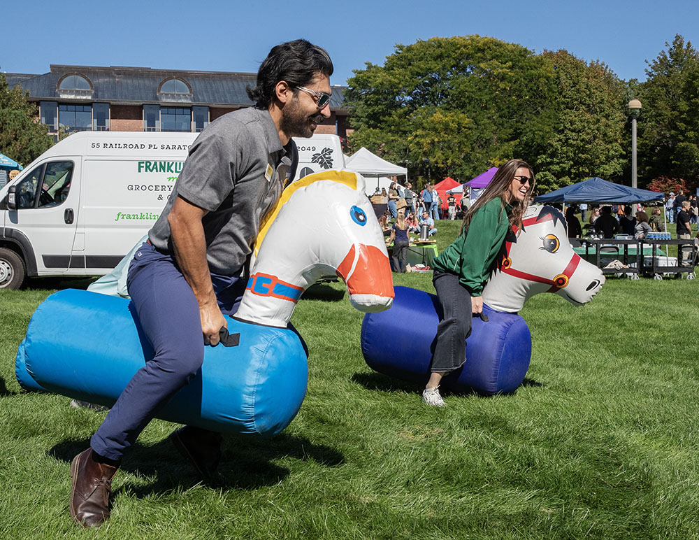 Two adults smile as they sit on blowup horses and race each other on a grass lawn. 