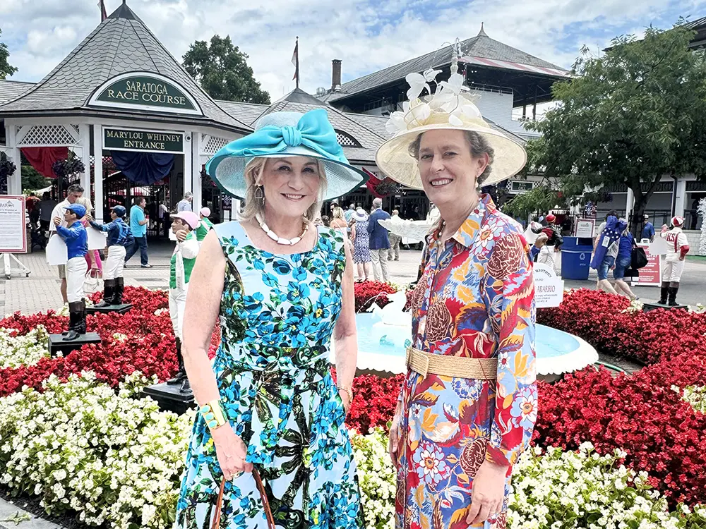 Two women wearing colorful dresses and hats in front of flowers at Saratoga Race Course