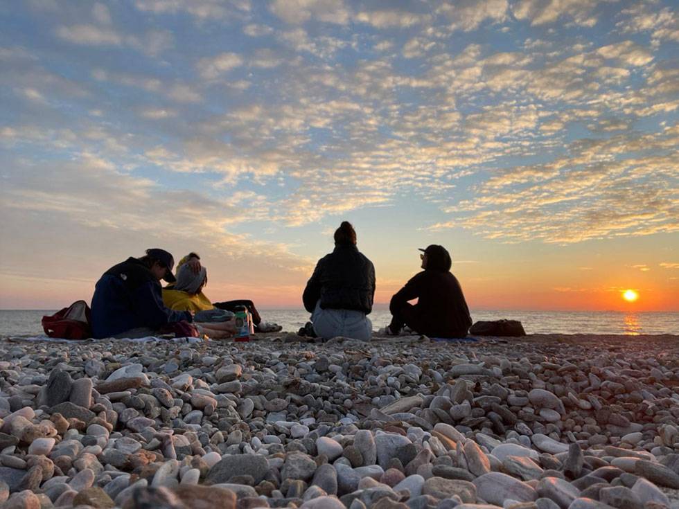 Four students sit on a rocky beach in Greece as the sun starts to set in the background.