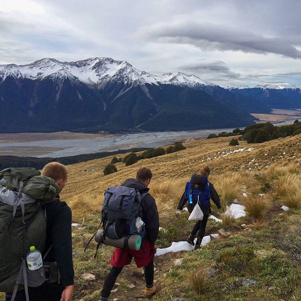 Students backpack through New Zealand back country with a lake a snow capped mountains in the background.