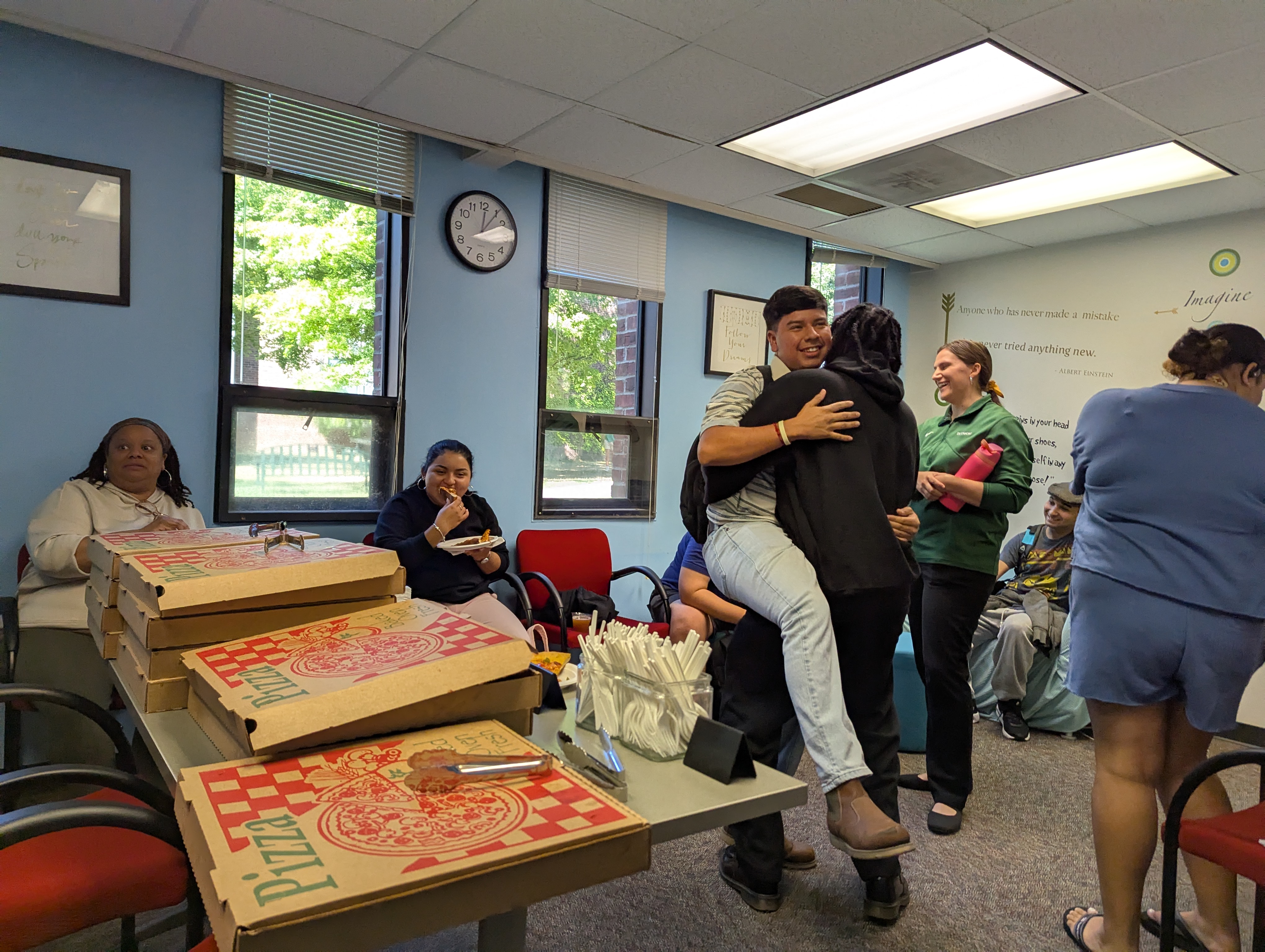 Students and staff gather in a campus lounge with pizza boxes on the table, sharing food, laughter, and warm hugs during a social event