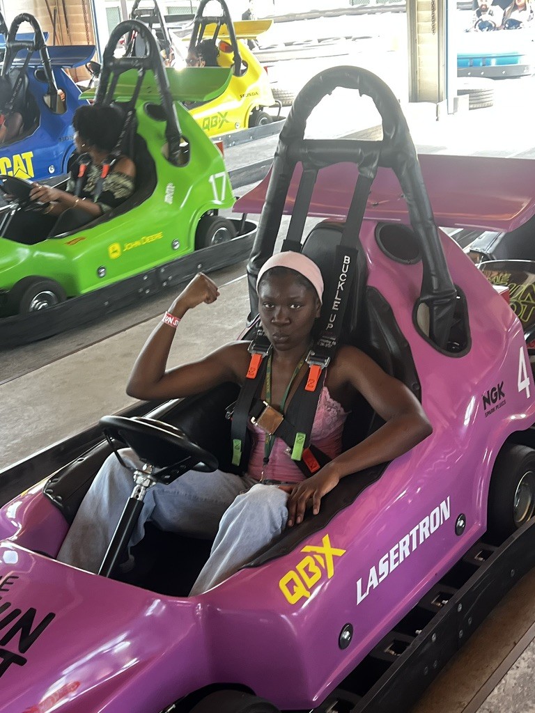 A student sits confidently in a bright purple go-kart, flexing her arm and smiling before a race at an indoor track.