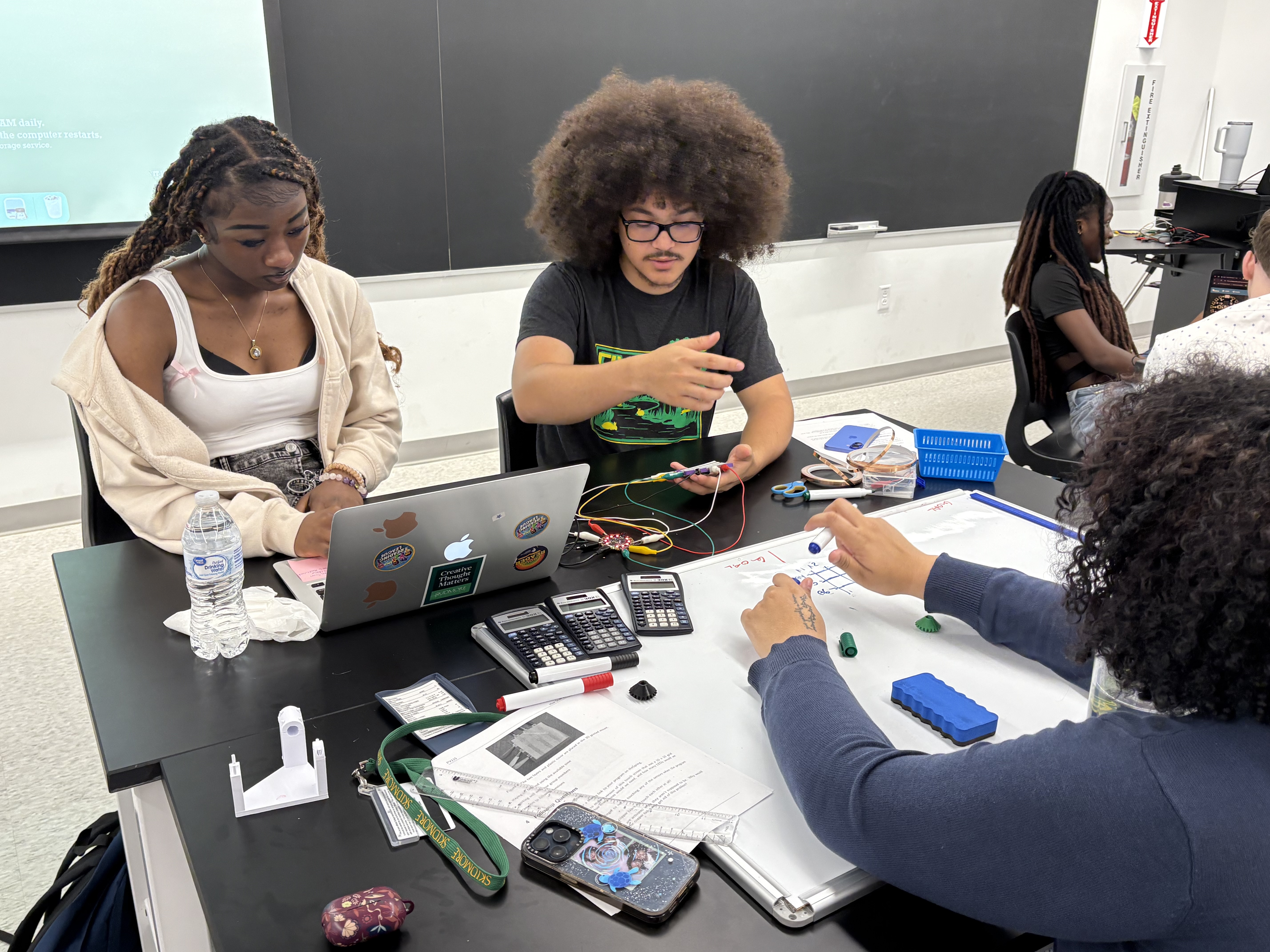 Students collaborate on a hands-on science or engineering project in a classroom, using laptops, calculators, and circuit components.