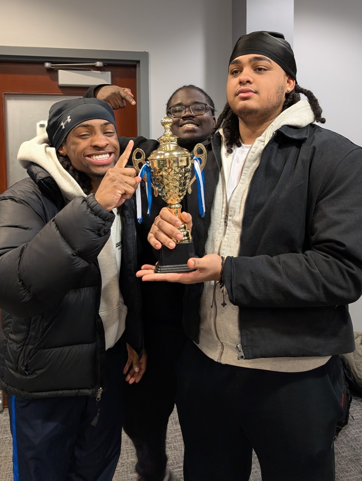 Three students smile and pose proudly with a gold trophy, celebrating their achievement indoors on campus.