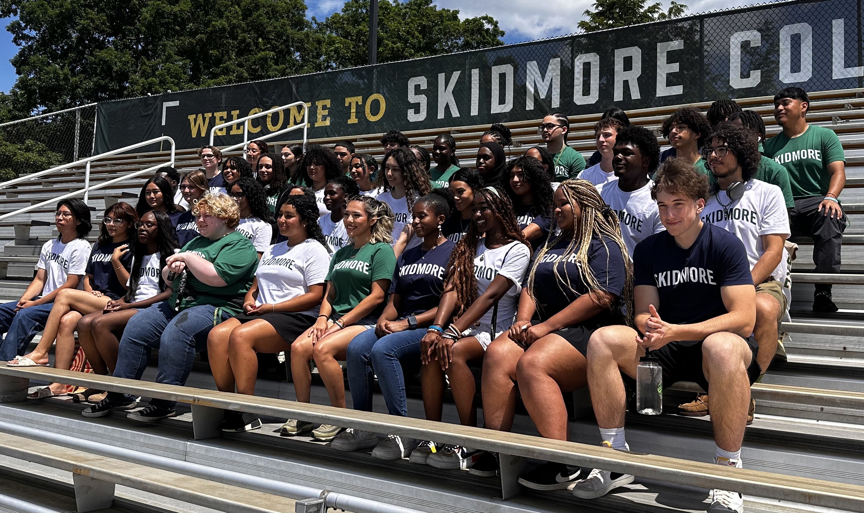 A diverse group of Skidmore College students sits on bleachers in Skidmore T-shirts, posing for a sunny group photo.