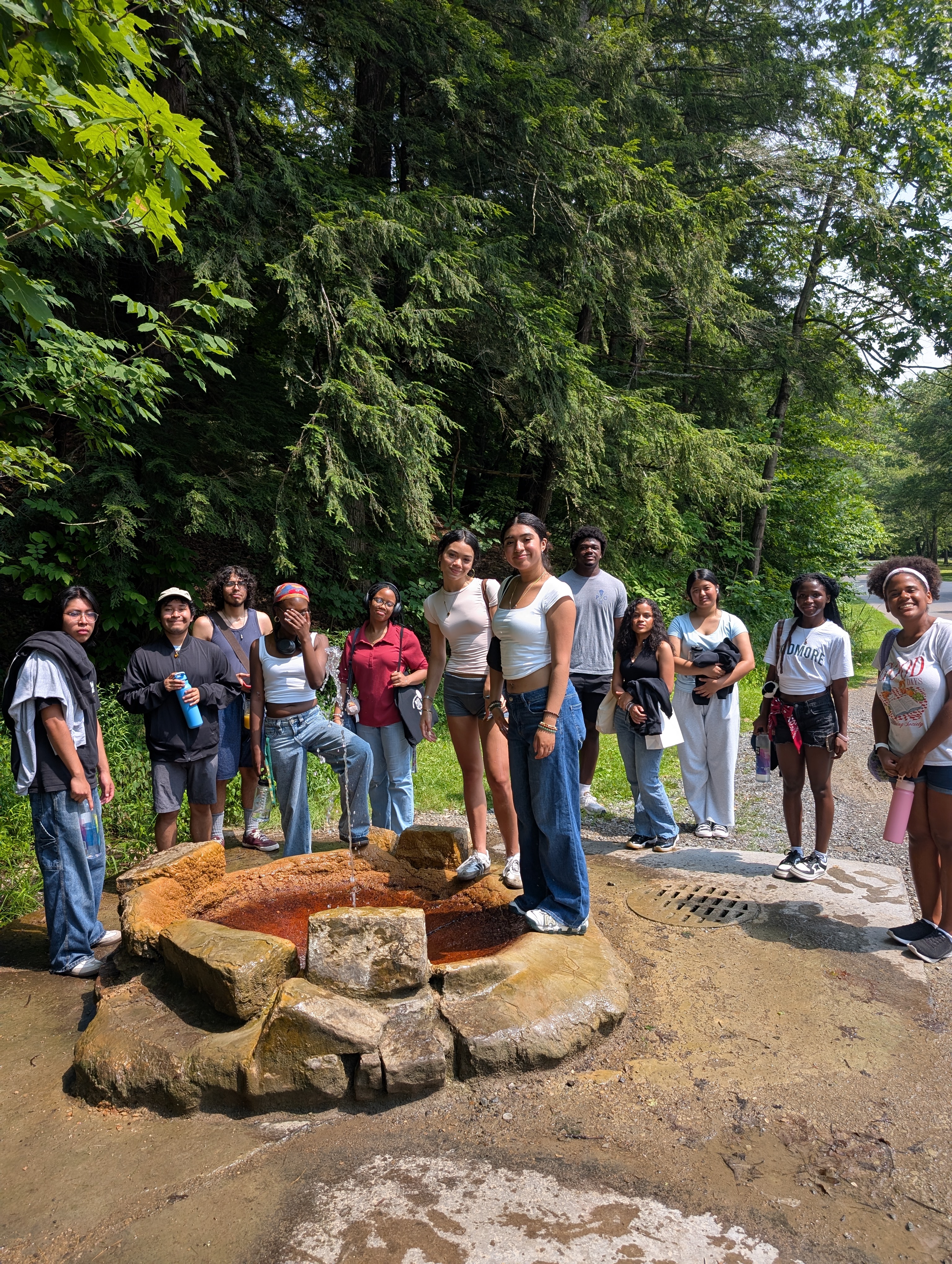 A group of students stands around a natural spring fountain in a wooded area on a sunny day, smiling for the photo.