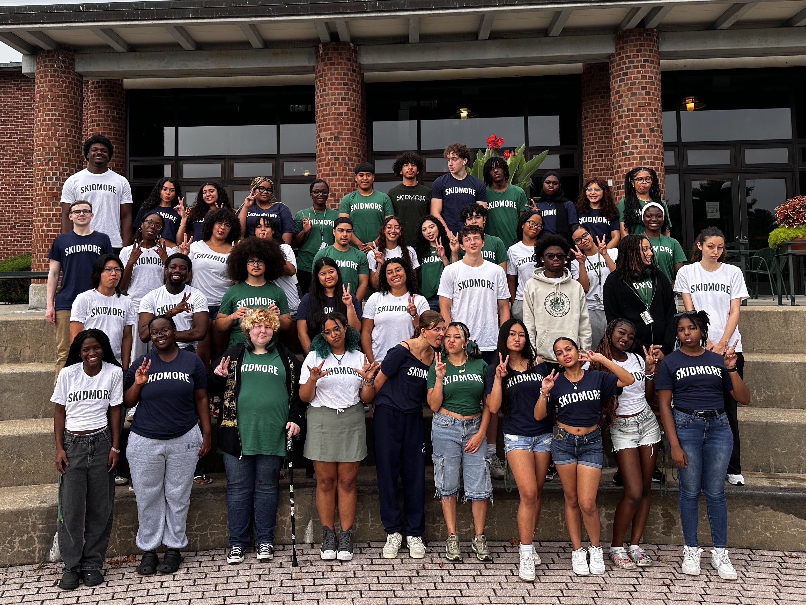 A large group of Skidmore College students wearing Skidmore T-shirts pose together on outdoor steps, smiling and making peace signs.