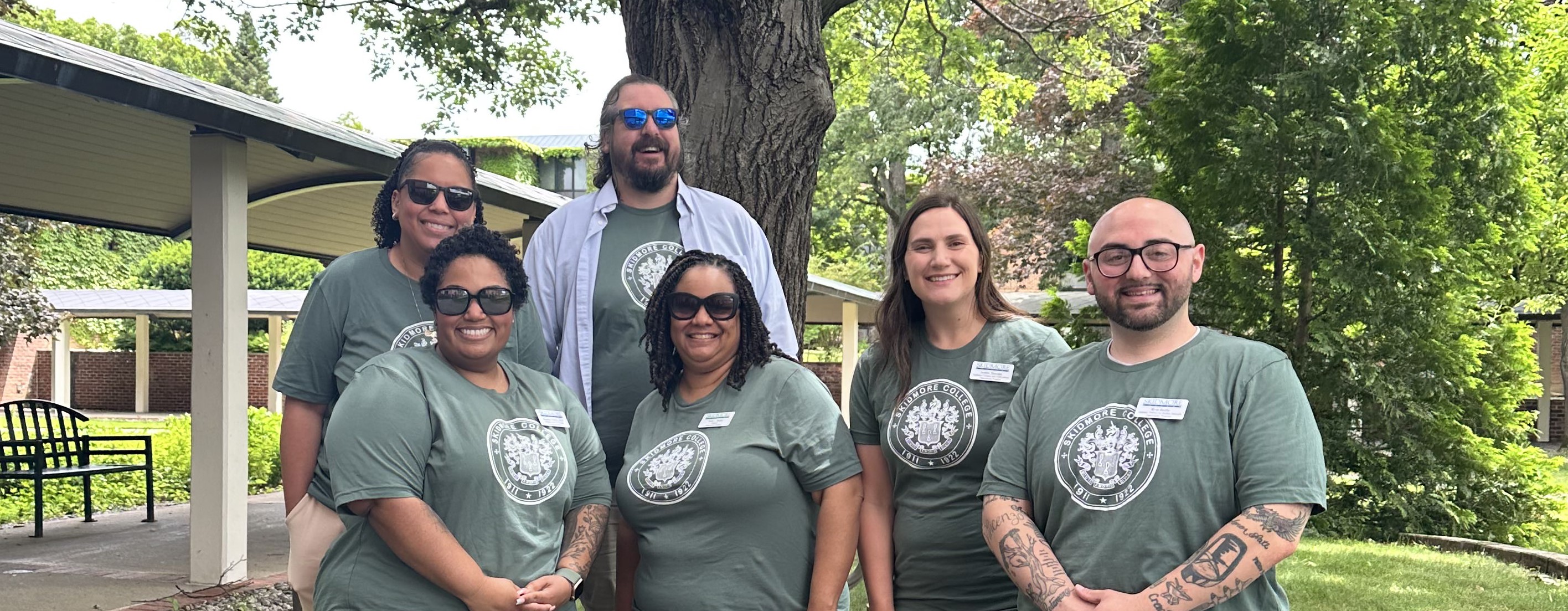 Six Skidmore College staff members wearing matching green Skidmore shirts smile together outdoors on campus.