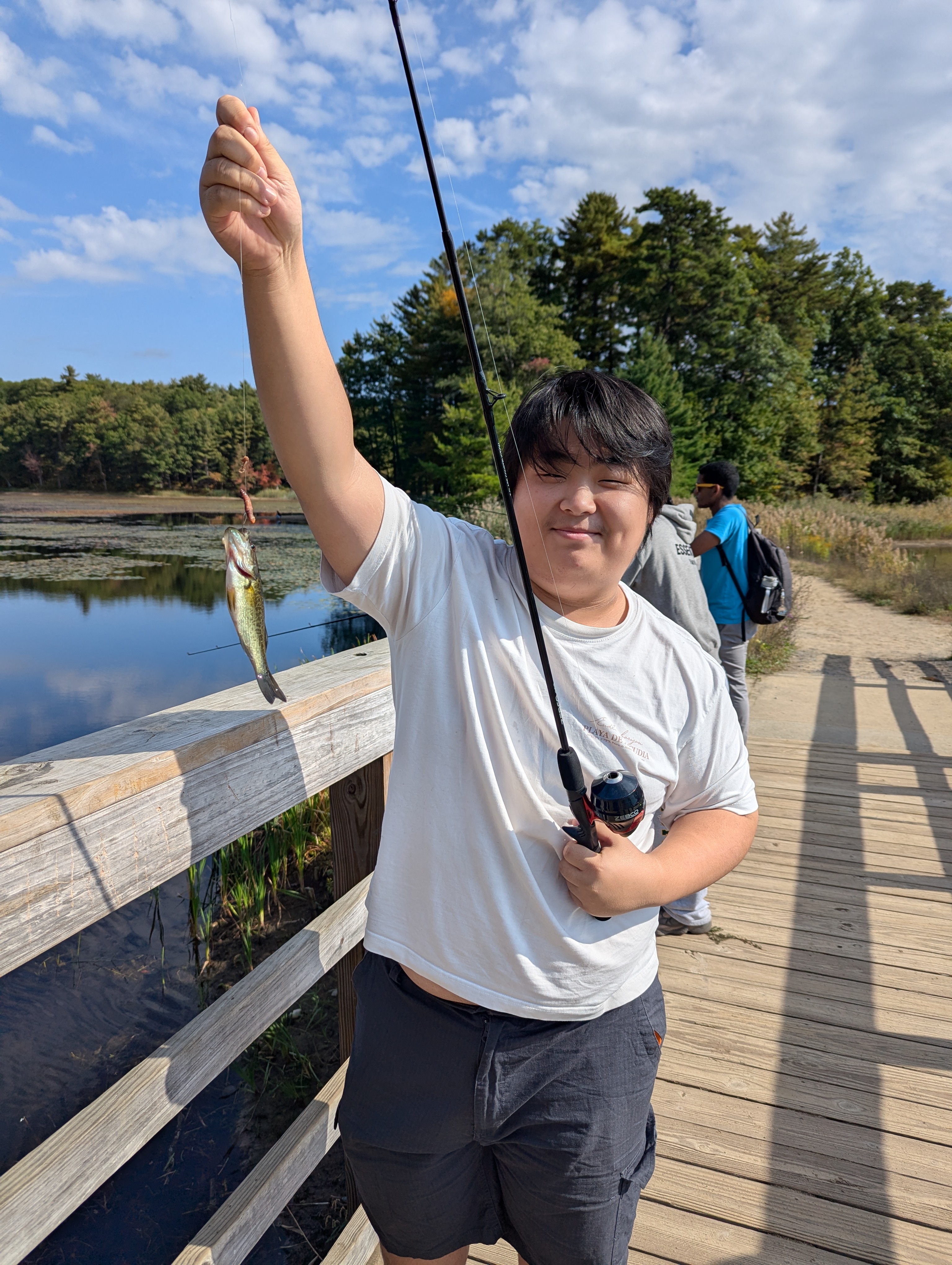 A student smiles while holding up a small fish caught on a fishing rod, standing on a wooden boardwalk.