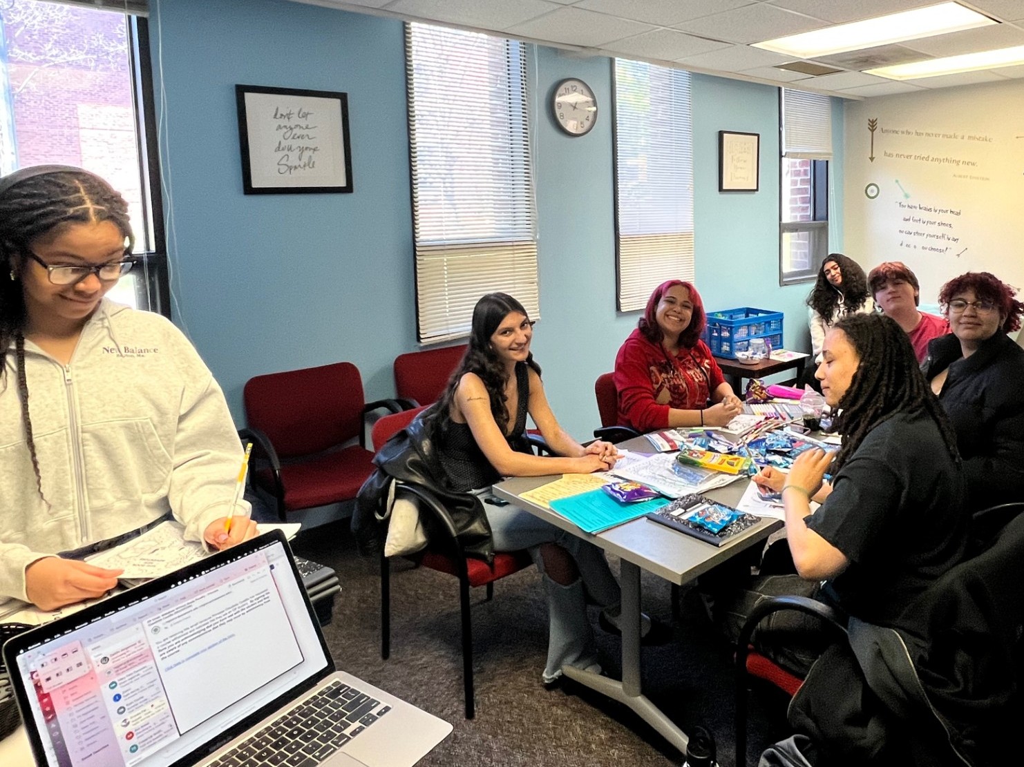 Students gather around a table in a campus office, smiling and working on writing and creative activities together.