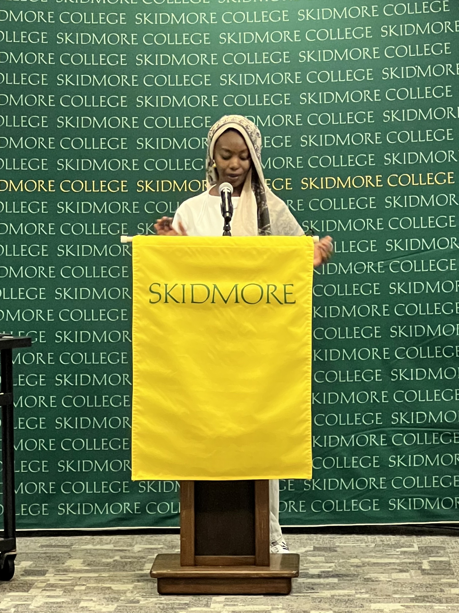 A student speaks at a podium with a Skidmore banner, addressing an audience during a campus event.