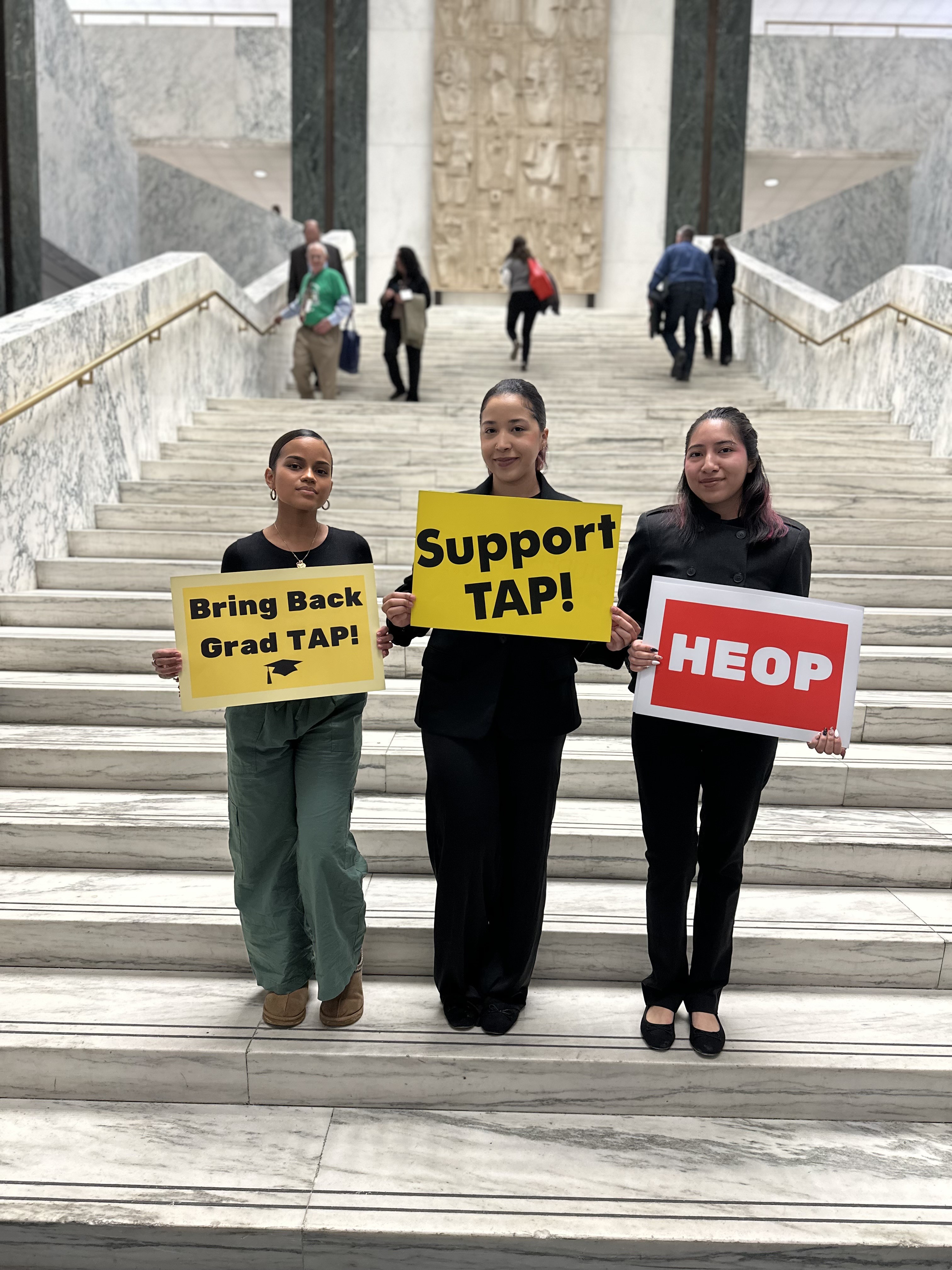 Three students stand on a marble staircase holding signs reading “Support TAP!,” “Bring Back Grad TAP!,” and “HEOP”