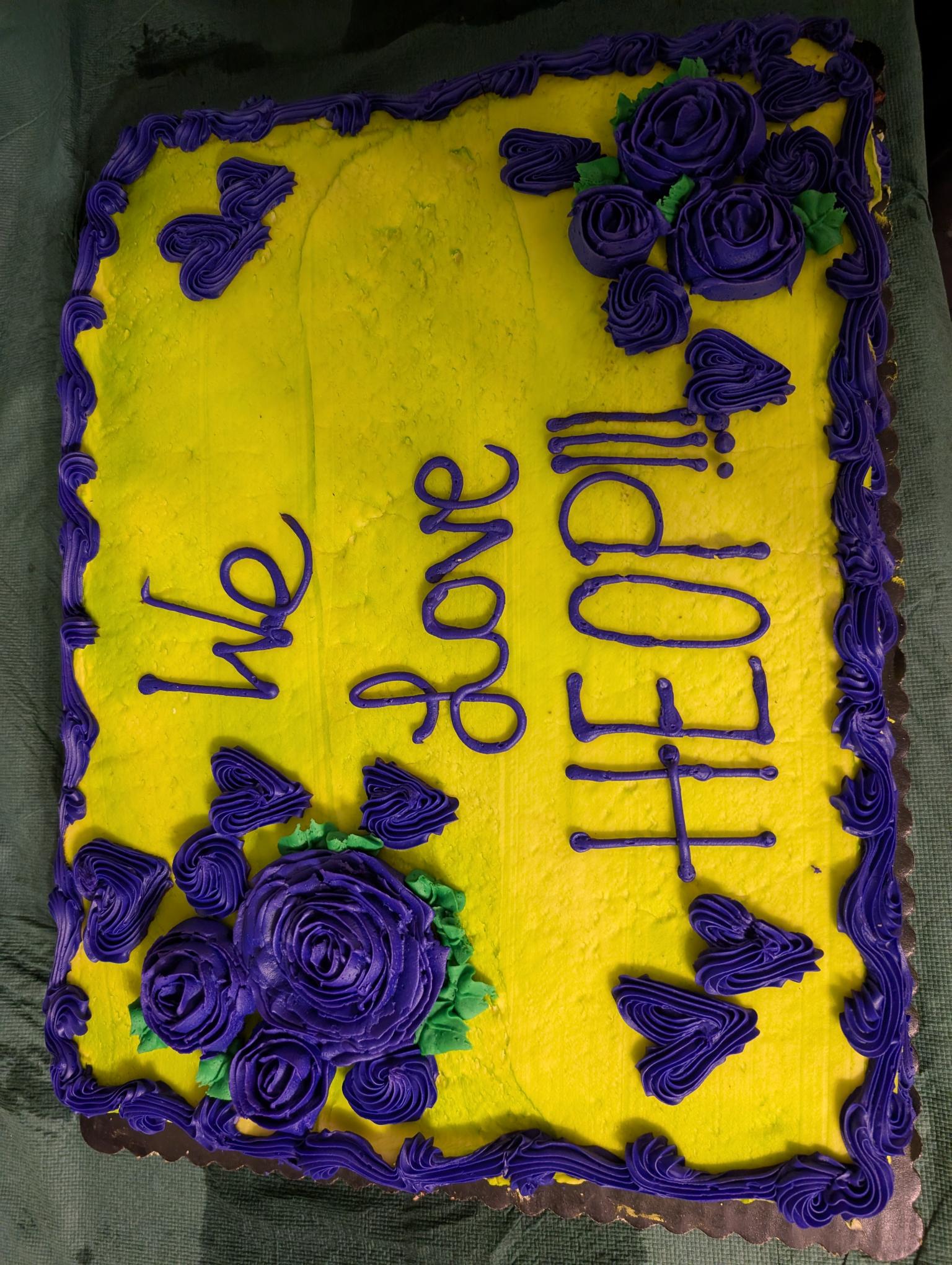 A decorated yellow cake with purple icing reads “We Love HEOP!!!,” 