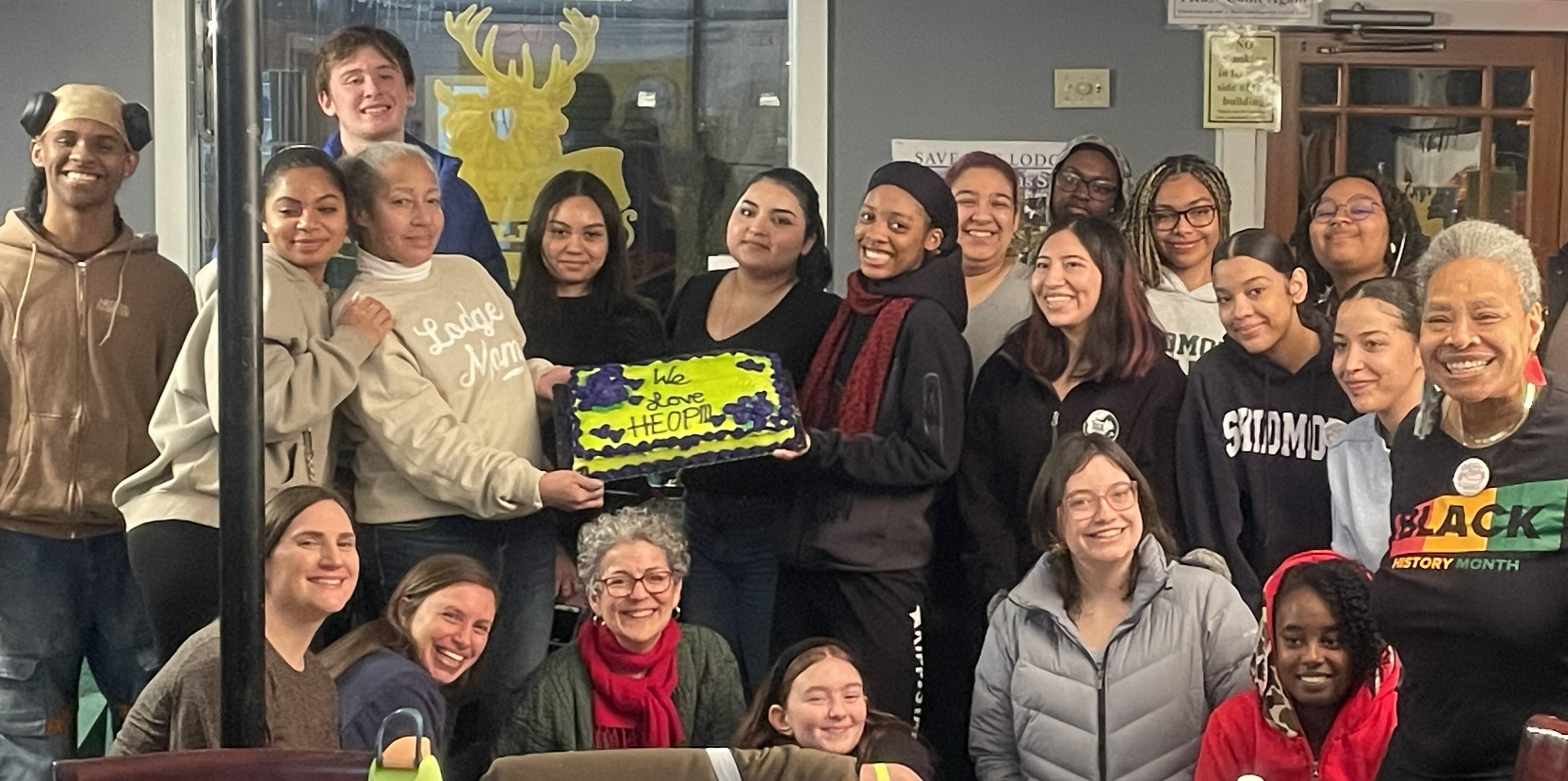 Group of diverse students and staff gathered indoors, smiling and posing together while holding a decorated cake that reads “We Love HEOP,”