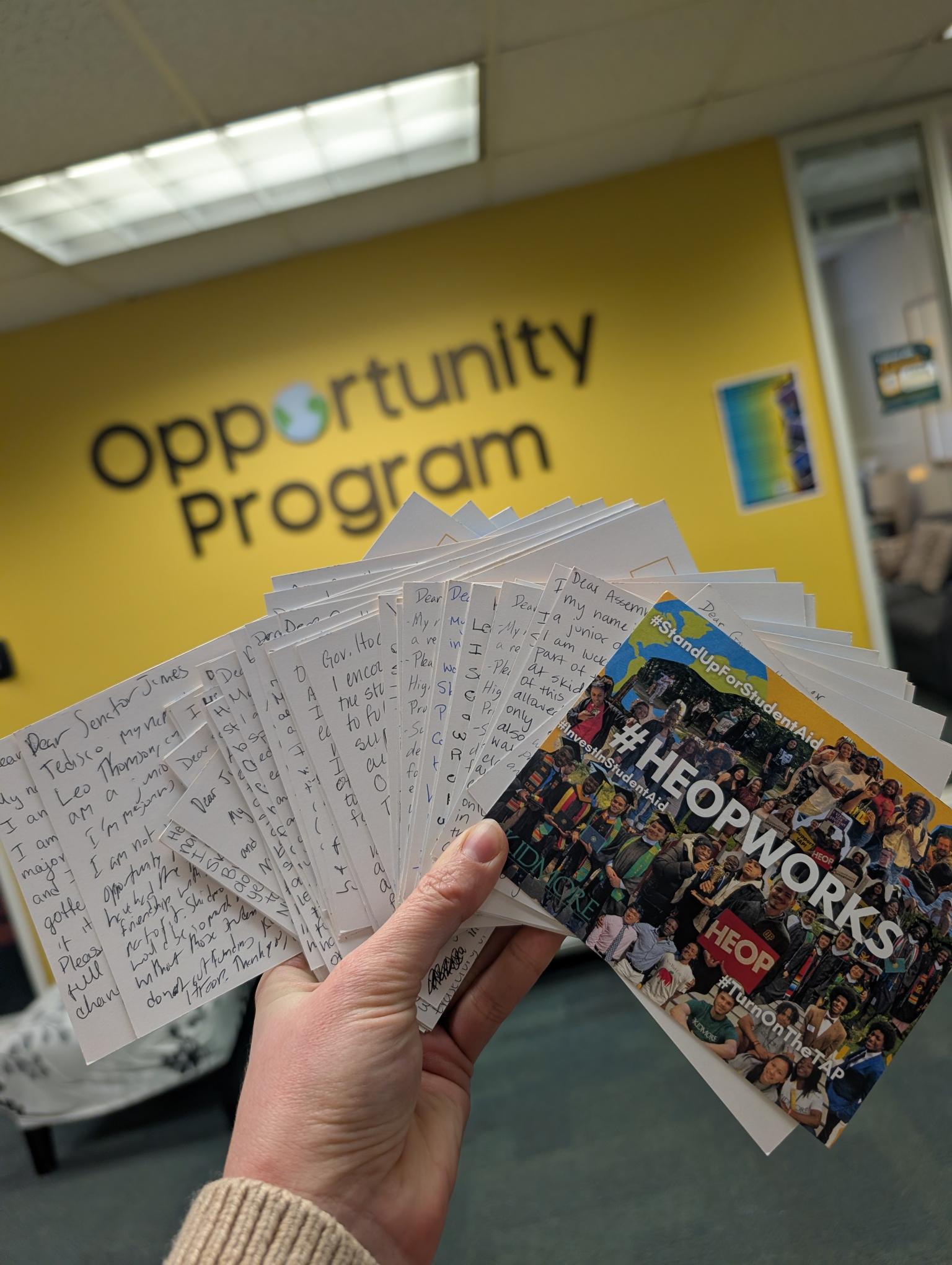A person holds handwritten letters in front of a yellow wall reading “Opportunity Program,” with a card showing students and “#HEOPWorks"