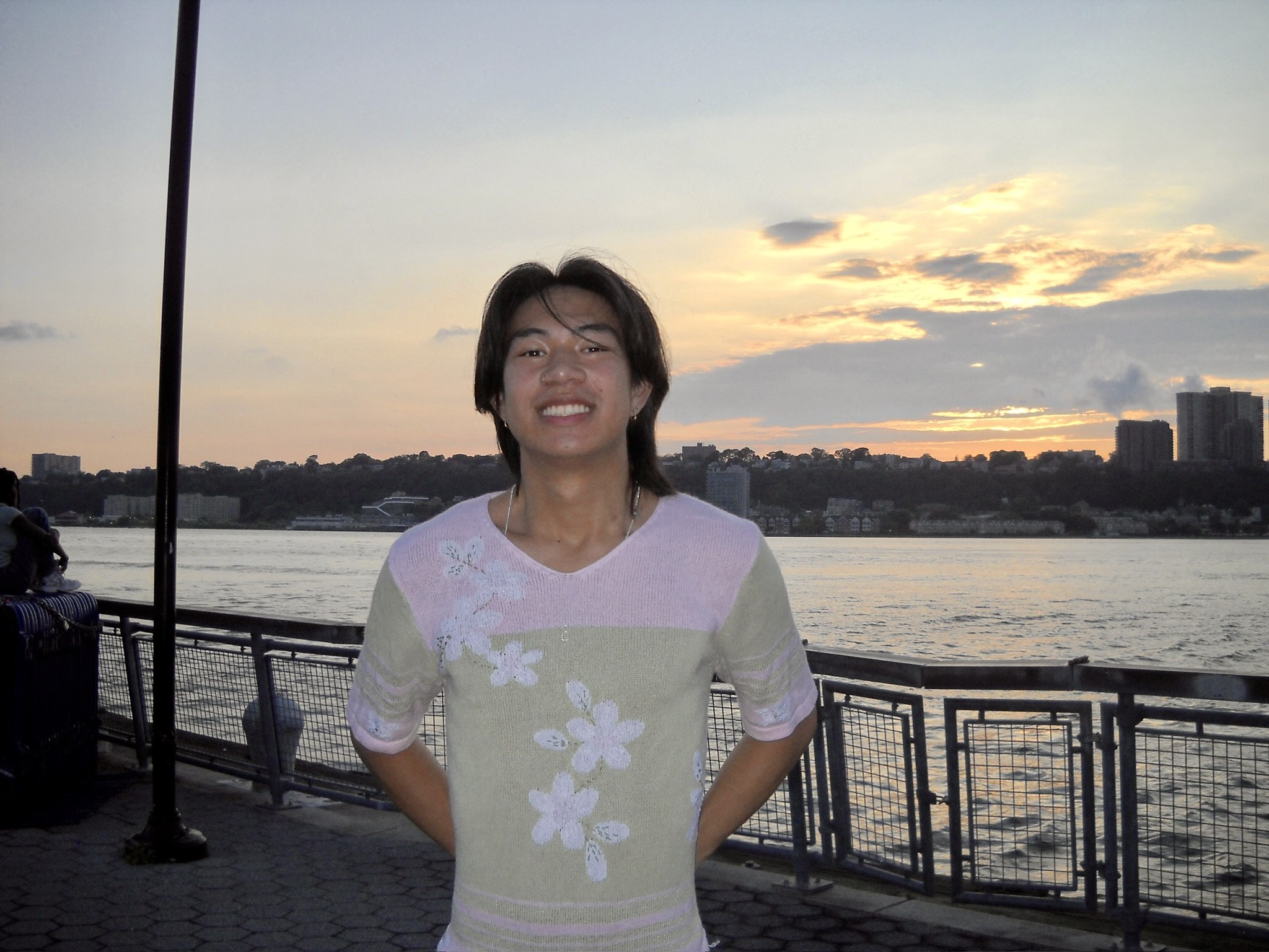 Photo os Asian male with shoulder-length black hair wearing a pastel pink and pastel green shirt with white flowers. He is outside in front of a river at sunset smiling.