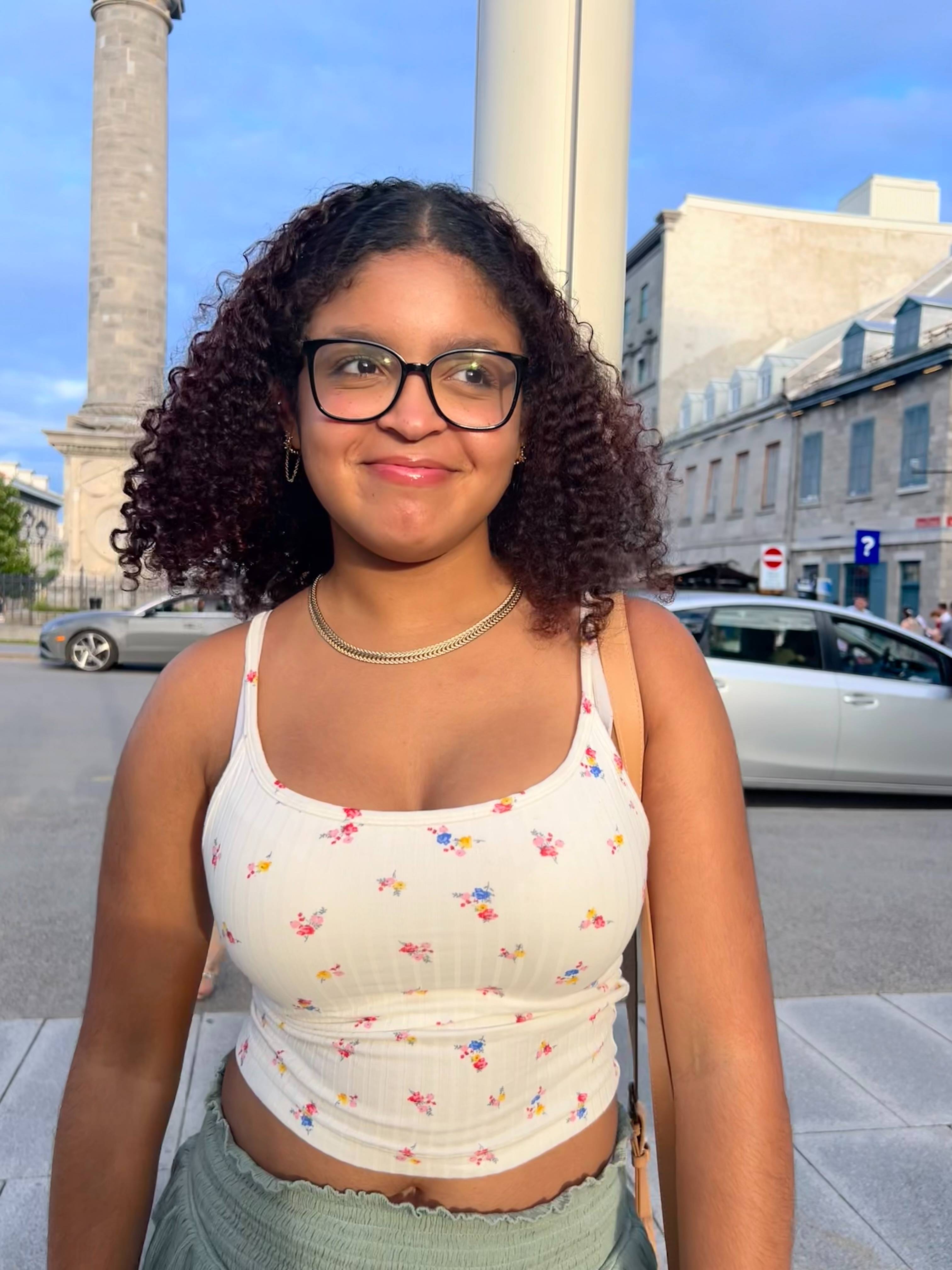 Photo of light-skinned Black tudent wearing glasses and white shirt with floral print. She is outside and smiling with a blue sky in the background