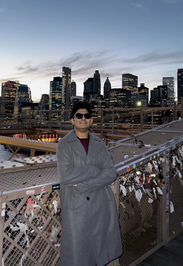 Photo of student staff wearing sunglasses in gray trenchcoat at dusk with city skyline in background