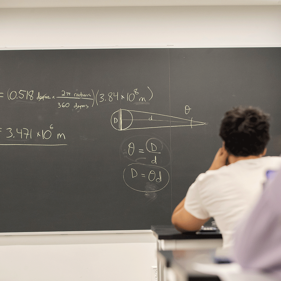 Student sits at a desk staring at a chalkboard with physics equations on a chalkboard