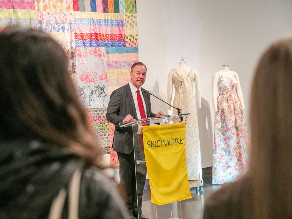 President Conner stands behind a Skidmore lectern in the Tang Museum, speaking to an audience, with a quilted piece of artwork and dresses in the background.