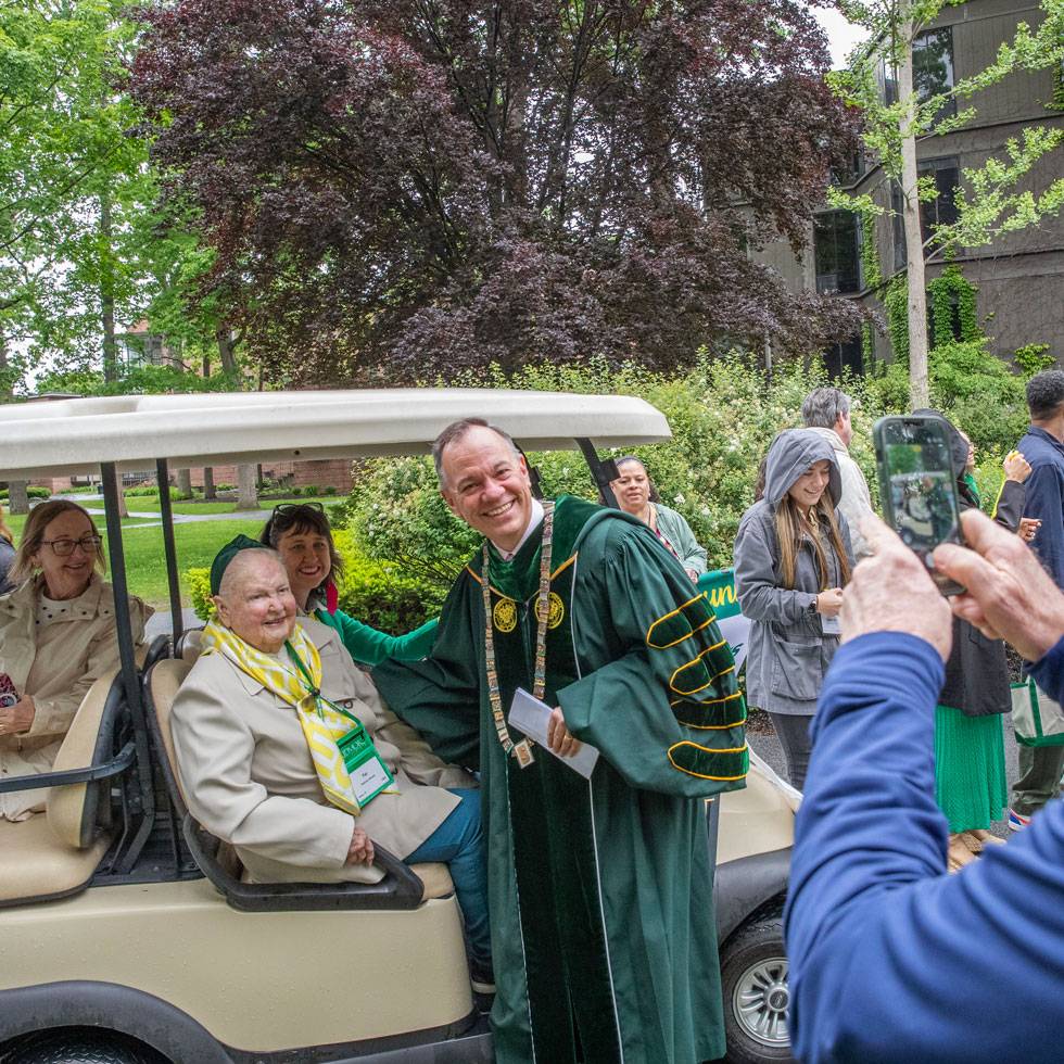 President Conner stands in Skidmore regalia taking a photo with alumni in a golf cart during reunion weekend.
