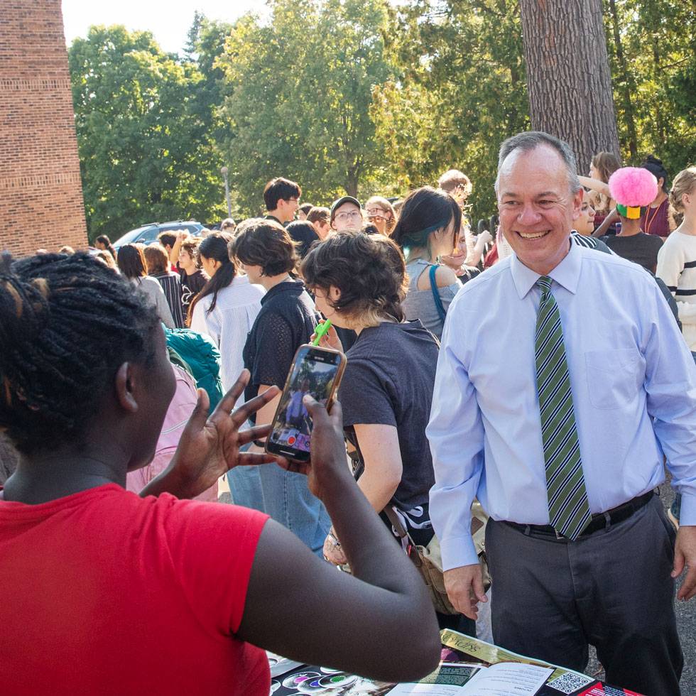 President Conner is smiling outside in a group of students, with one student in the forefront taking his photo with their iphone.