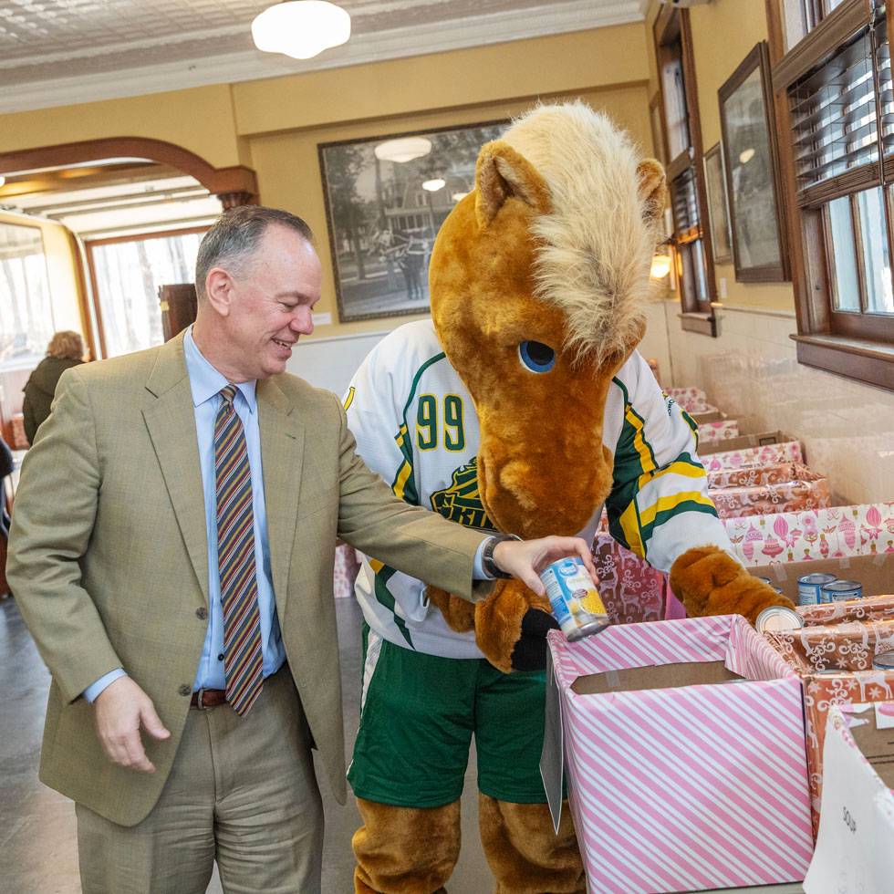 President Conner stands with Skidmore's thoroughbred mascot putting cans of food in wrapped box.