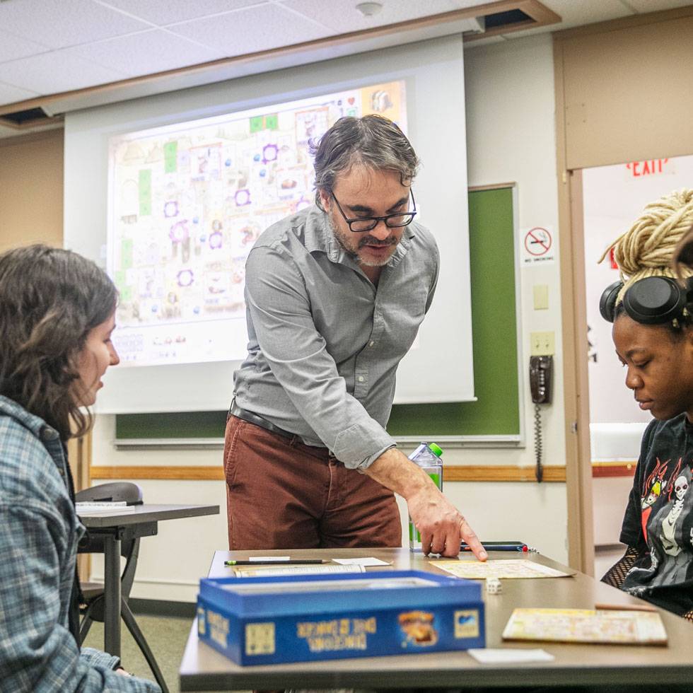 Professor Hassan Lopez works with two students in a classroom. He is leaning over a table they are sitting at, pointing to a game they are working on, to provide instruction. 