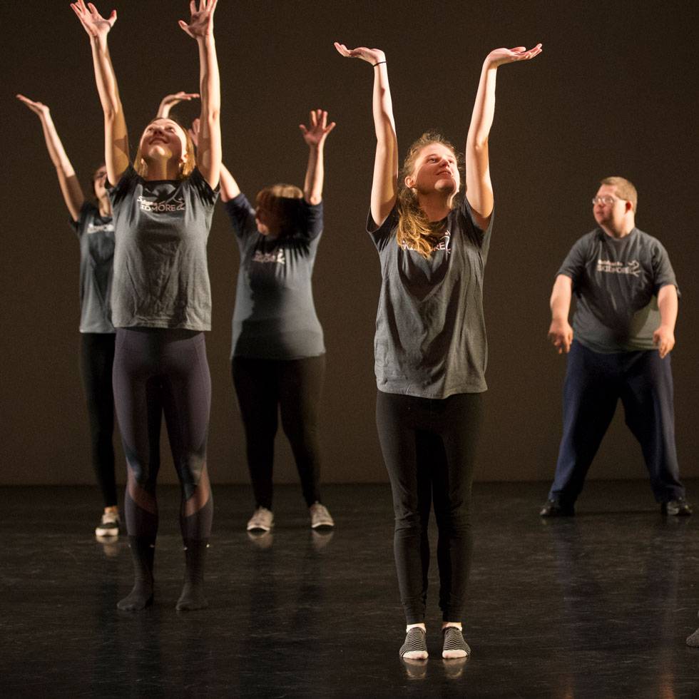 Skidmore students and patients of Saratoga Bridges dance on a dark stage with their arms thrown in the air and wearing matching grey t-shirts.