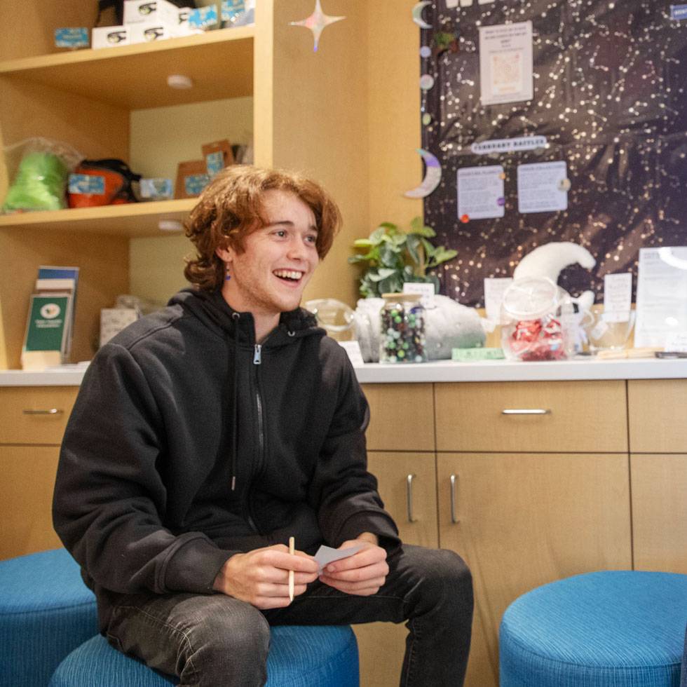 Vojta Kubala sits in health services on a teal ottoman smiling and holding a pen and piece of paper in their hand.