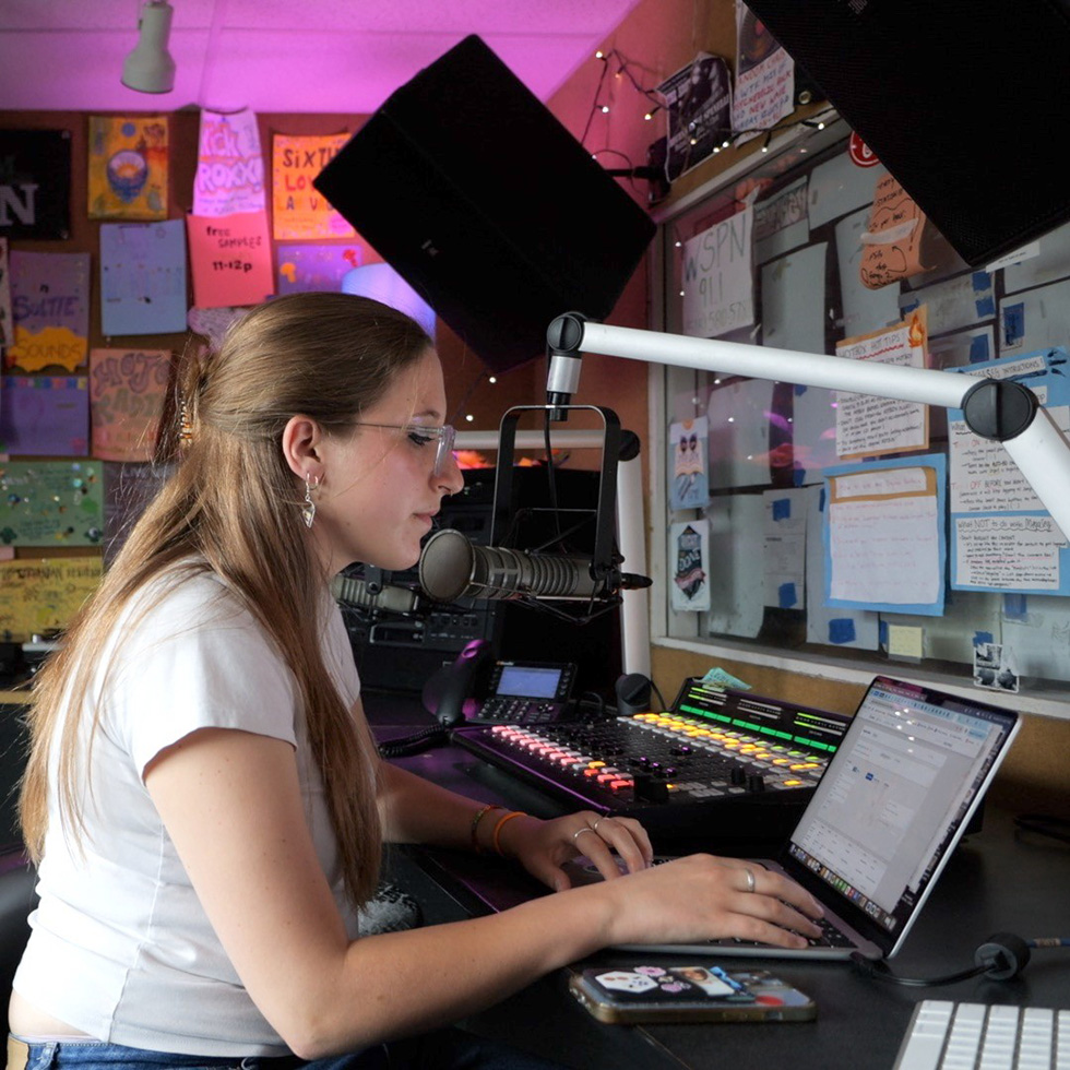 Oli Landsman sits in a radio recording studio working on a laptop with a pink light overhead.