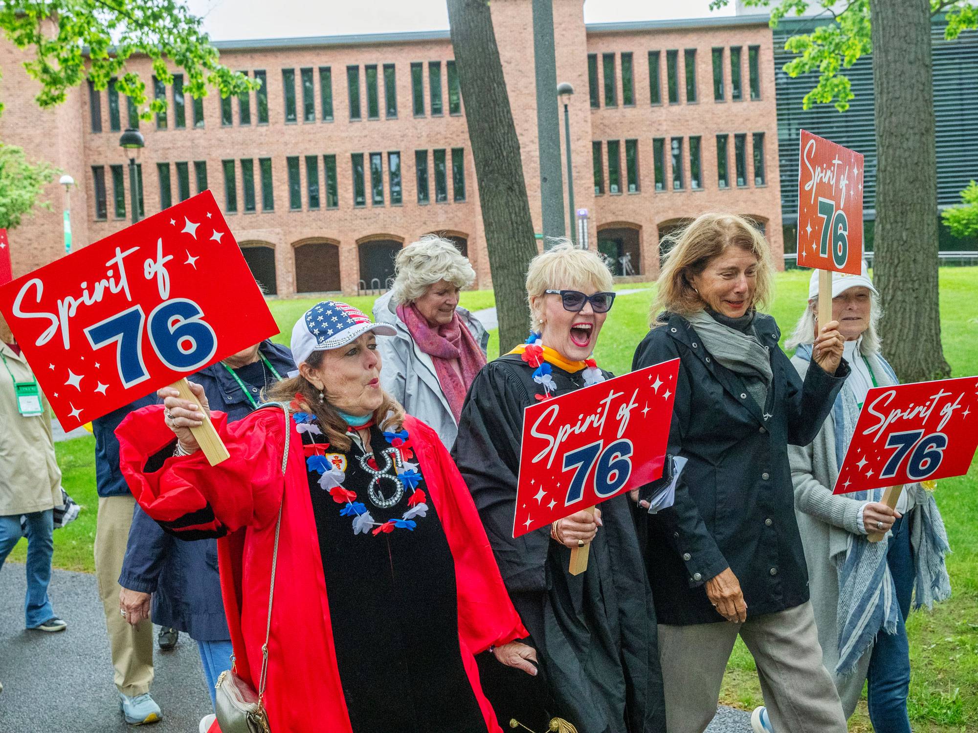 A group of classmates from the class of 1976 walk outside in the reunion parade holding bright red sides that say "the Spirit of '76"