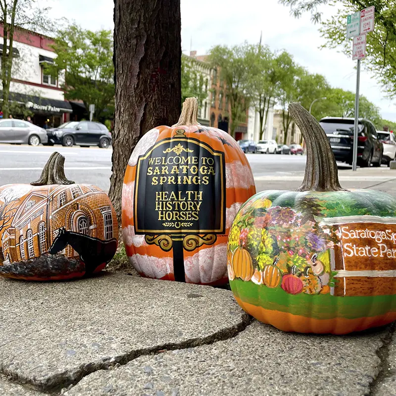Hand-painted pumpkins sit on a Saratoga Springs sidewalk, each showcasing local icons like historic buildings, fall harvest scenes, and the city’s “Health, History, Horses” welcome sign — part of a festive downtown display.