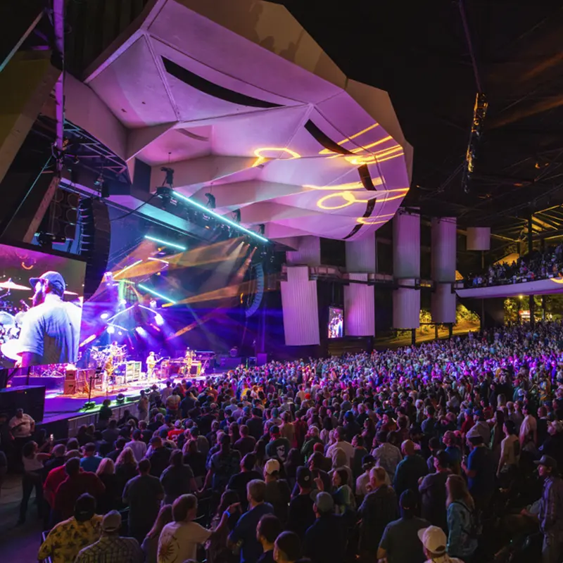 A packed crowd enjoys a high-energy concert at Saratoga Performing Arts Center, lit by colorful stage lights under SPAC’s distinctive open-air amphitheater.
