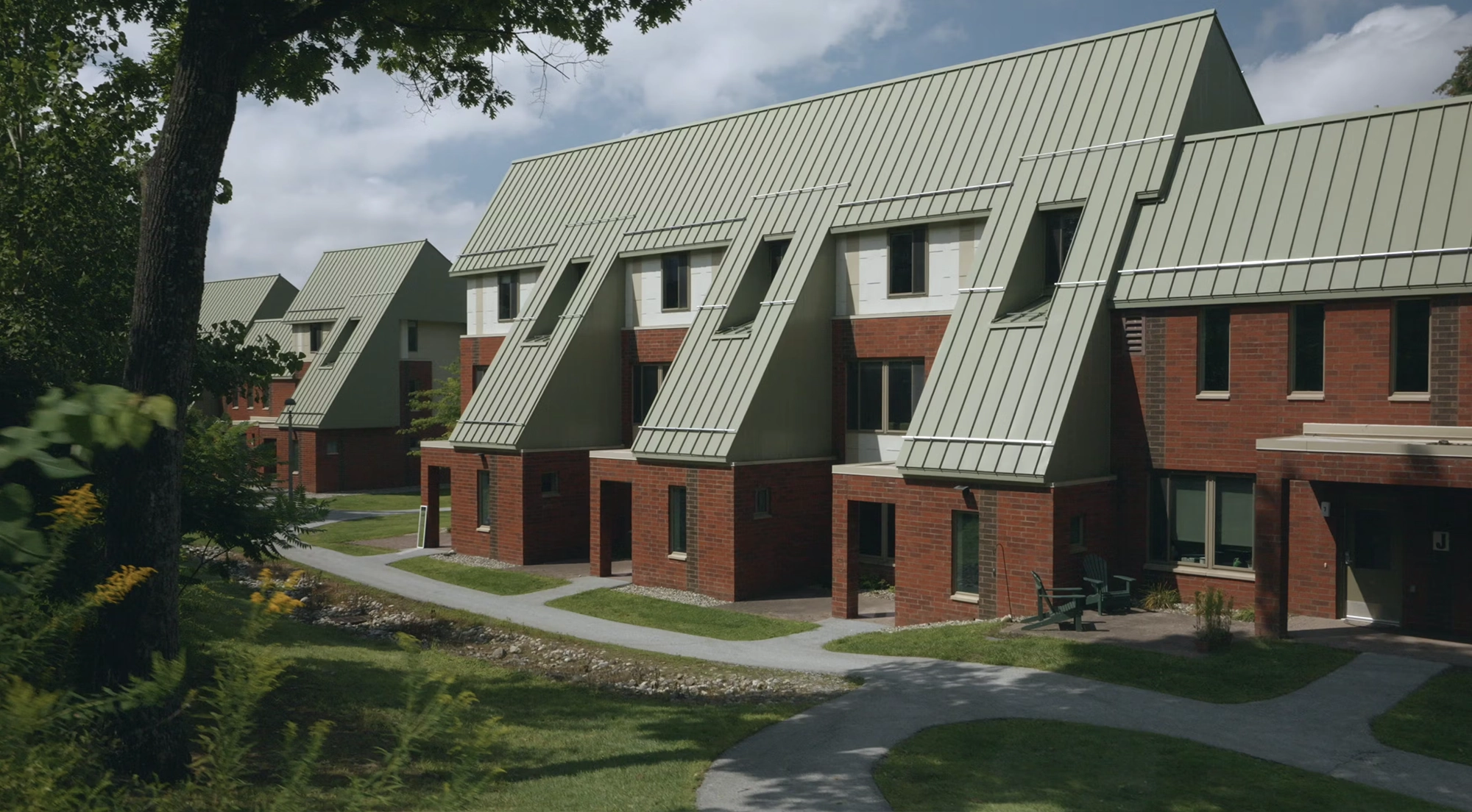 Row of modern red-brick residence halls at Skidmore College, featuring distinctive green metal roofs with large angular dormer windows; the buildings are surrounded by trees, landscaped paths, and green lawns under a partly cloudy sky.