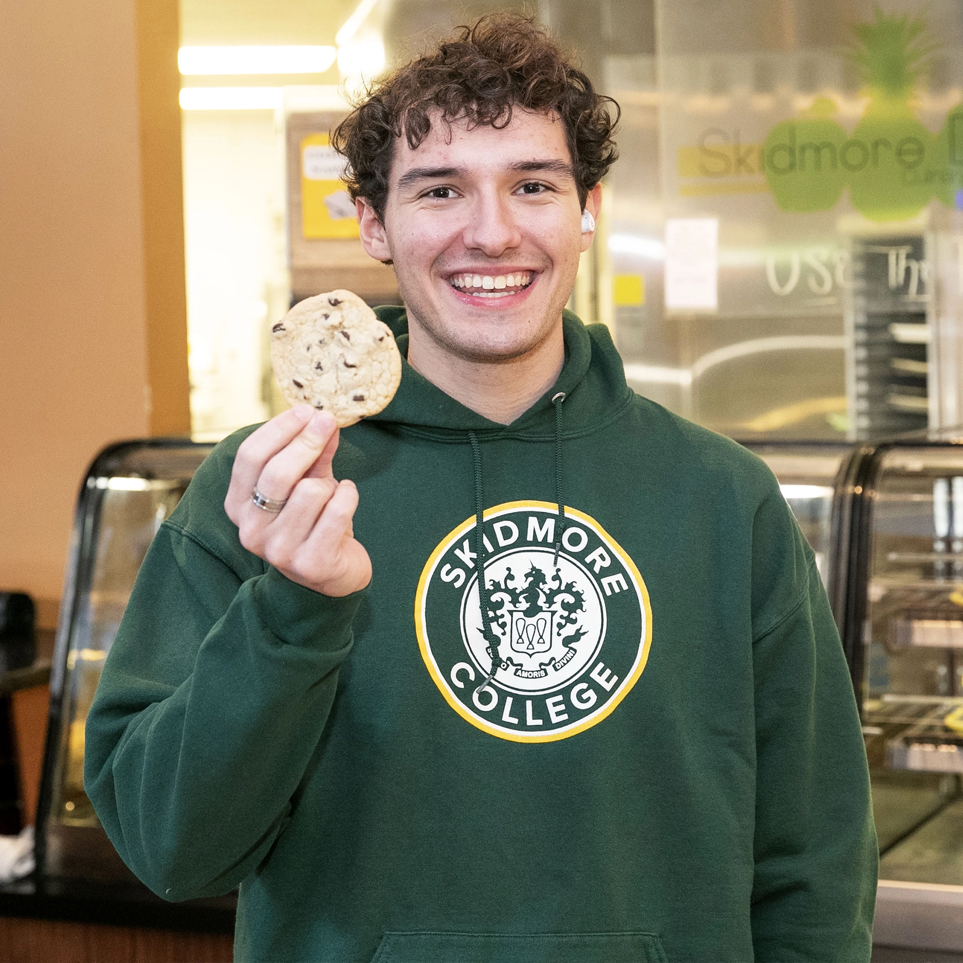 Smiling Skidmore student in a campus dining hall holding up a chocolate chip cookie while wearing a green Skidmore College hoodie.er