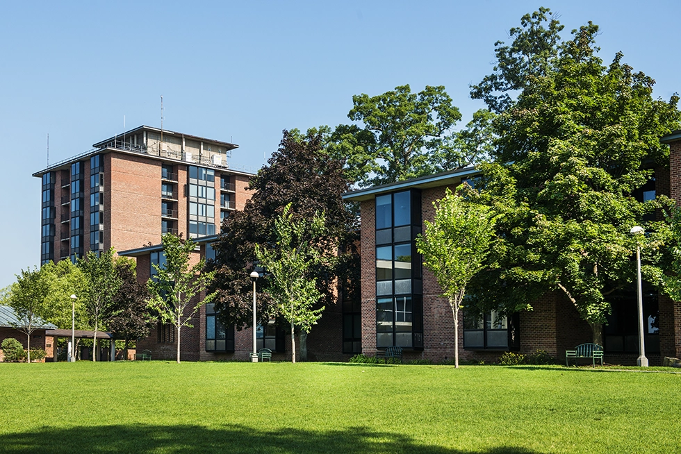 Skidmore College residence halls with red brick exteriors and tall windows overlook a sunny green quad lined with trees and benches.
